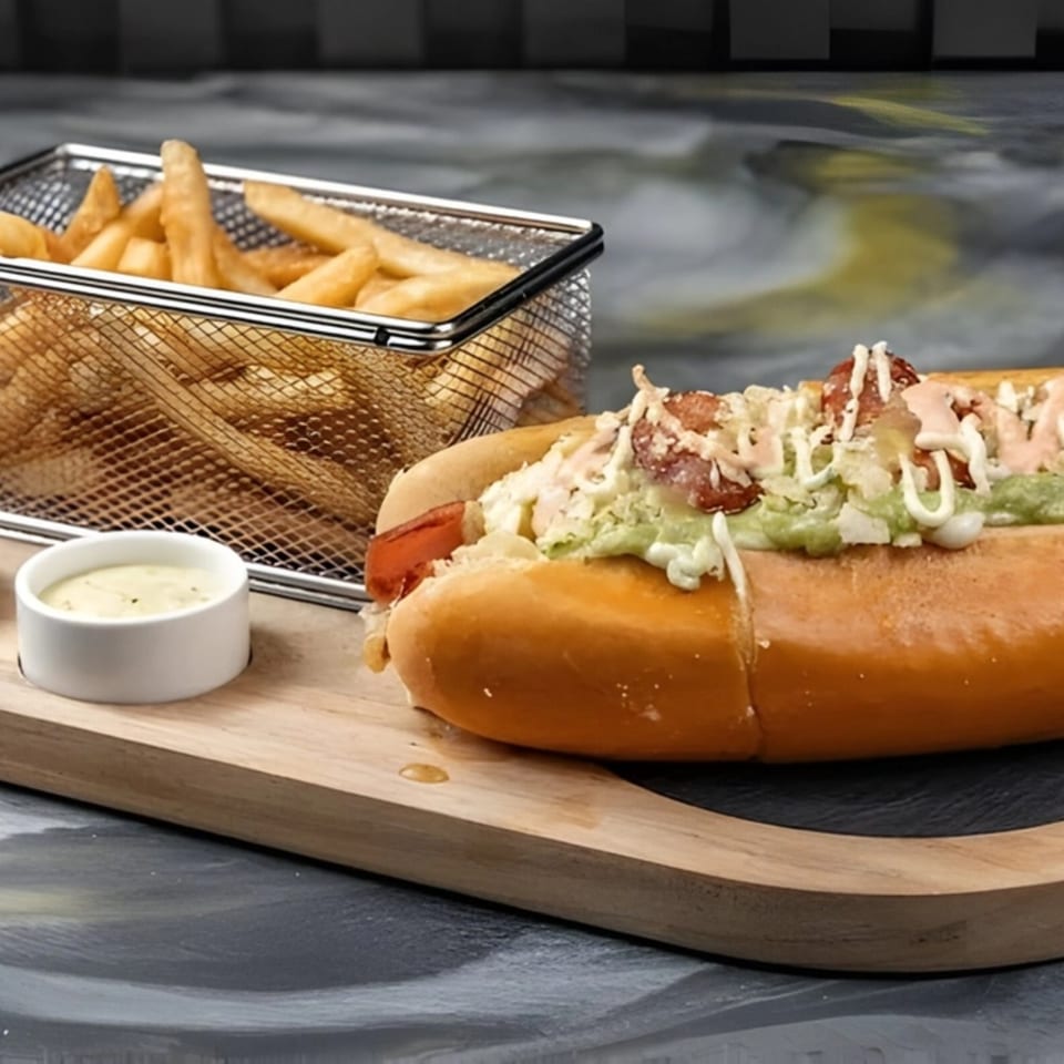 A Colombian hot dog with toppings, a side of fries in a metal basket, and a dipping sauce.